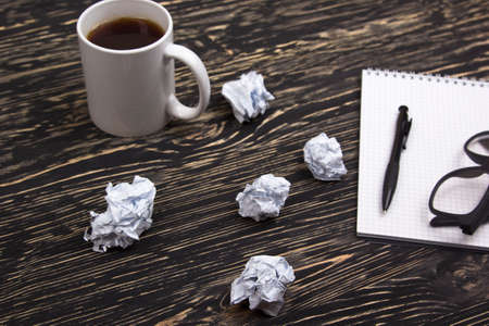 Crumpled paper balls with eye glasses mug, pen and notebook on wooden deskの写真素材