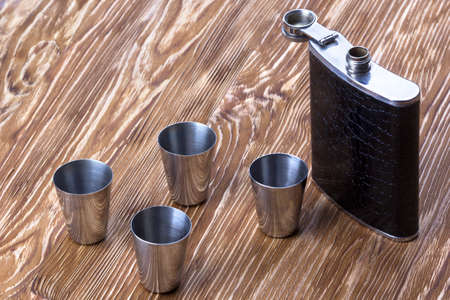 Metal flask trimmed leather and four metallic, sturdy shot glasses on a wooden backgroundの写真素材