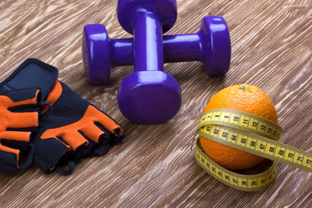 Dumbbells, orange, fitness gloves and measuring tape on wooden backgroundの写真素材