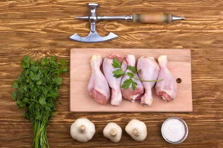 raw chicken drumstick on the board with salt, garlic, parsley and ax on wooden tableの写真素材