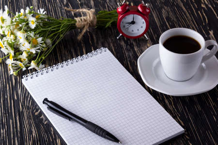 blank notebook, alarm clock, charmomile and coffee cup on wooden background.の写真素材