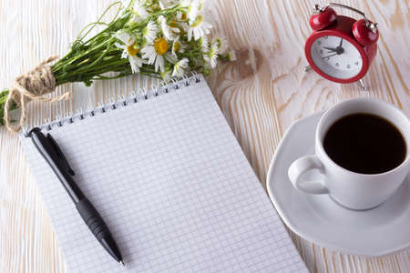 blank notebook, alarm clock, charmomile and coffee cup on wooden background.