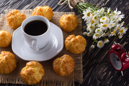 Eclair, coffee cup, alarm clock and charmomile on wooden background.