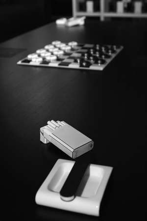 White ashtray, pack of cigarettes, box of matches and chessboard on a black table in an home...