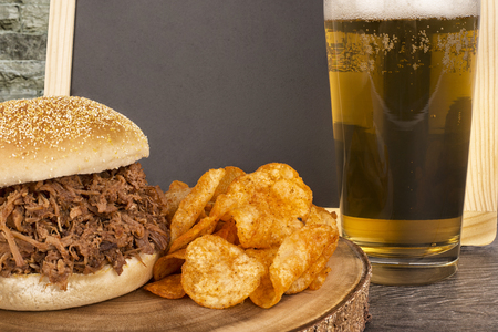 Close up on pulled pork sandwich and blonde beer on wooden plank, empty blackboard in background.