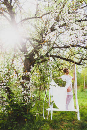 Young bride with blond hair in white negligee and stockings posing on a rope swingの写真素材
