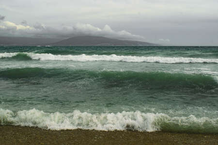 waves of sea foam are running along the pebble beach, mountains covered with clouds are visible in the backgroundの写真素材
