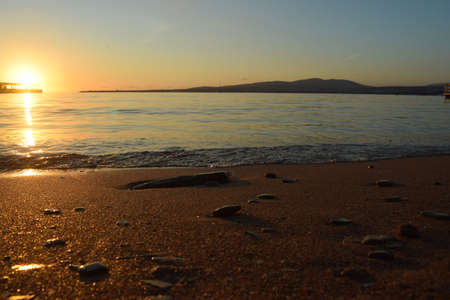 the sun setting behind the horizon, sandy dull seashore strewn with stones and lazy waves running onto the beachの写真素材