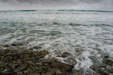 autumn stormy sea, waves on the coastal rocky beach, water jets leaving the sea after the surf, a ship on the horizon and clouds over the seaの写真素材