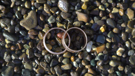Two gold wedding rings lying on beach stones shining on sun light close up macro. Transfusion of light on rings. ocean wave washed on themの写真素材