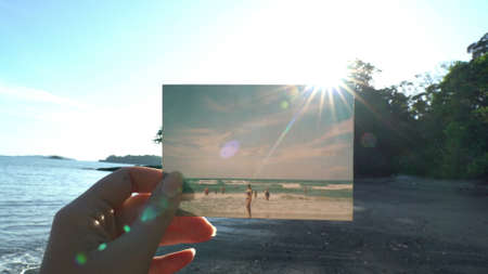 Close up hand holding photograph in front of beautiful tropical beach travel concept. Sun shining beautiful day inspirational landscape, sunbeam raysの写真素材