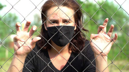 woman in a medical protective mask behind a metal wire fence. The concept of social distance and quarantine during the coronavirus pandemicの写真素材