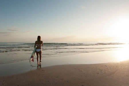 Young attractive surfer girl on beach with surfboard at sunrise