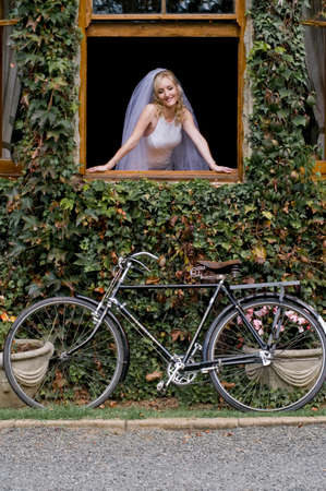 Young beautiful blonde bride looking out window frame with vintage bicycle outdoorsの写真素材