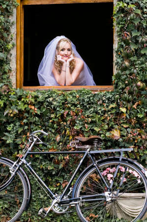 Young beautiful blonde bride looking out window frame with vintage bicycle outdoorsの写真素材
