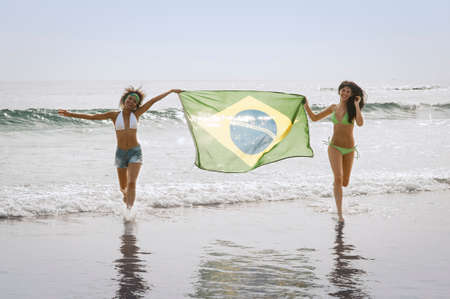 Two attractive girls running along beach with Brazil flagの写真素材
