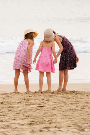 3 sisters stand on beachfront facing the ocean.Whispering in sisters ear