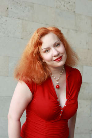 Young red-haired woman in red shirt posing against grey limestone wallの写真素材