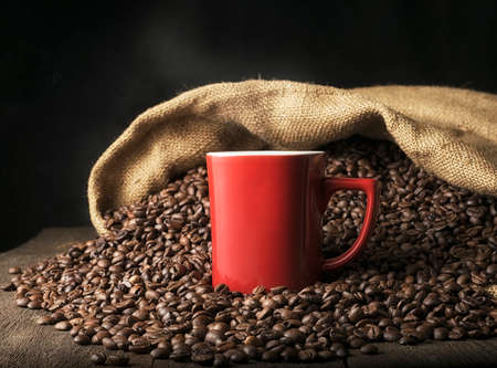 White mug with hot steaming coffee on the background of a heap of coffee beans spilled from a bag on the tableの写真素材