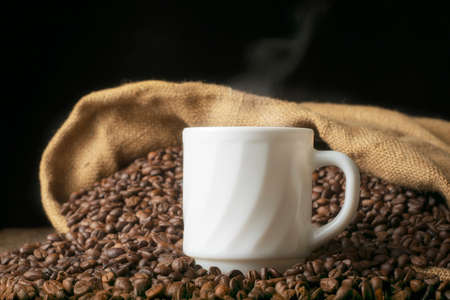 White mug with hot coffee on the background of a heap of coffee beans spilled from a bag on the tableの写真素材