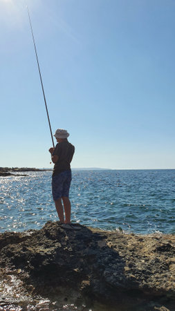 Fisherman with fishing rod standing on rock and looking at seaの写真素材
