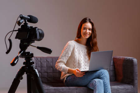 Young teen girl blogger makes a video stream on the couch at home. A video camera on a tripod and a laptop.の写真素材