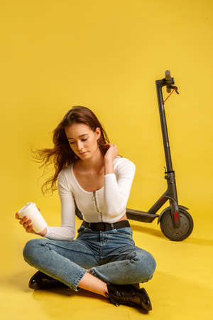 Young beautiful girl with electric scooter and coffee cup. Yellow background. Technology and freedom of movement.の写真素材