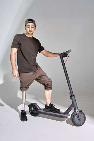 Disabled young man with leg prosthesis with Kick scooter on white background. Sun light.