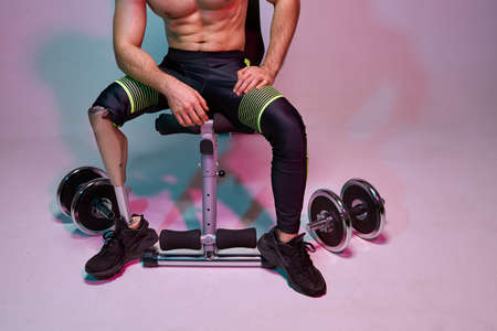 A young muscular man with a disability with a prosthetic leg is training in a gym with dumbbells....