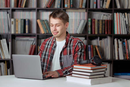Happy guy listens to a remote audio lesson in the library with wireless headphones and prints a report on a laptop.の写真素材