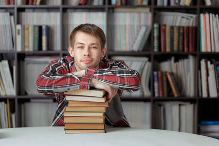 Cool guy-student with a smile sitting at the library table and hugging a stack of books, background...
