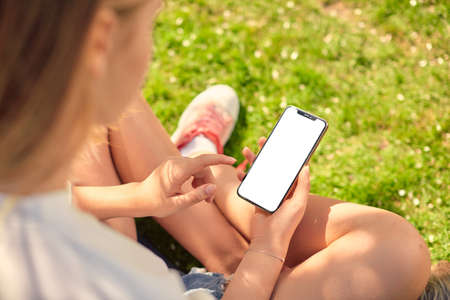 Cute teen girl looks at the screen of her smartphone sitting on a green lawn in the garden. Close-up phone display.の写真素材