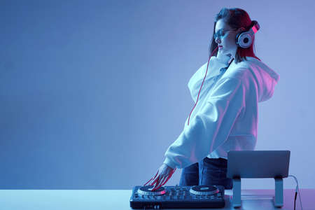 Cool young girl DJ mixes music on a mixing console and laptop, in stylish clothes, glasses on a neon background.の写真素材
