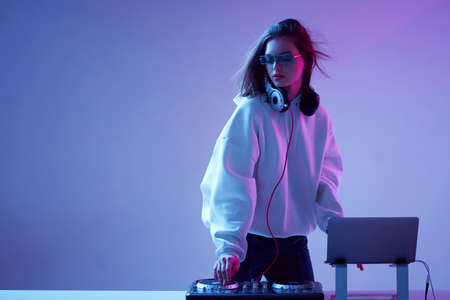 Cool young girl DJ mixes music on a mixing console and laptop, in stylish clothes, glasses on a neon background.の写真素材