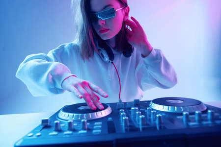 Cool young girl DJ mixes music on a mixing console and headphones, in stylish clothes, glasses on a neon background.の写真素材