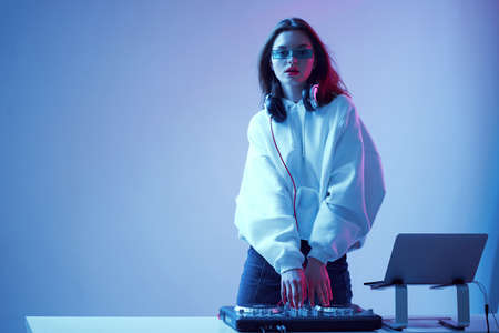Cool young girl DJ mixes music on a mixing console and laptop, in stylish clothes, glasses on a neon background.の写真素材
