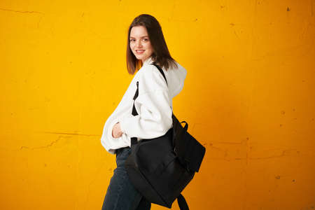 Portrait of a young pretty girl student in a white hoodie, casual clothes, with a bag, against a concrete orange wall.の写真素材