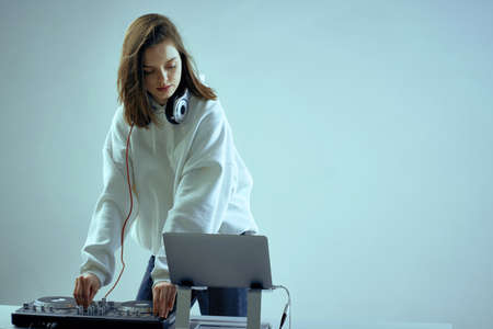 Cool young girl DJ mixes music on a mixing console and laptop, in stylish clothes, on white blue background.の写真素材