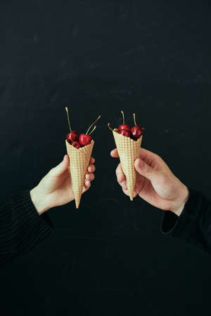 A girls hand and a mans hand hold waffle cones filled with fresh cherries on a black background.の写真素材