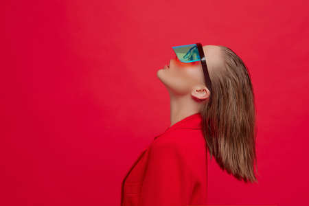 A cool stylish girl with a fashionable hairstyle and stylish glasses with a large glass poses on a bright red backgroundの写真素材