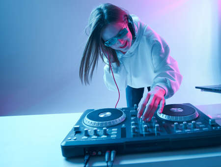 Cool young girl DJ mixes music on a mixing console and laptop, in stylish clothes, glasses on a neon...