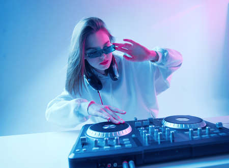 Cool young girl DJ mixes music on a mixing console and headphones, in stylish clothes, glasses on a neon background.の写真素材