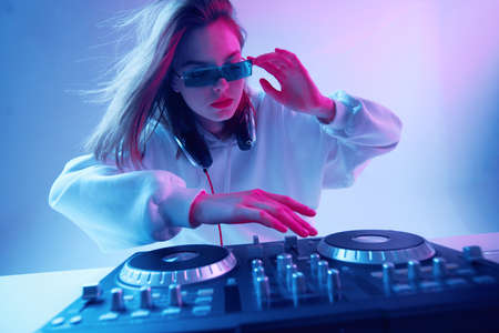 Cool young girl DJ mixes music on a mixing console and headphones, in stylish clothes, glasses on a neon background.の写真素材