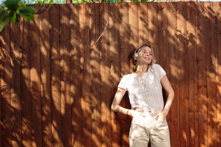 Portrait of a pretty girl in wireless headphones, standing against a wooden fence in the backyard on a Sunny day.の写真素材