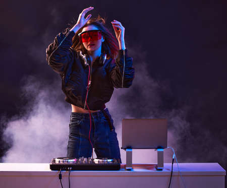 Stylish girl DJ in glasses headphones and a bomber mixes music dancing at a party in the neon light in the smoke .の写真素材
