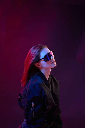 Portrait of a cool girl in black bomber jacket with sunglasses on a dark background in neon light.