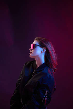 Portrait of a cool girl in black bomber jacket with sunglasses on a dark background in neon light.の写真素材