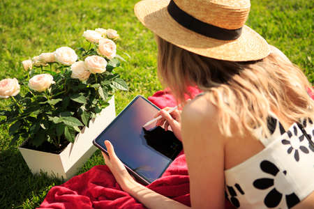 Young pretty girl in hat Illustrator draws remotely on tablet on green lawn with roses, photo for...