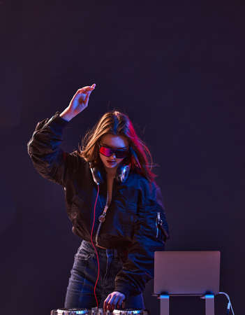 Stylish DJ girl with headphones and glasses on the bomber jacket mixes the music at the party and neon lights.の写真素材