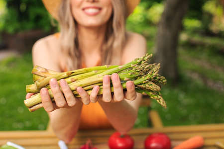 Young pretty girl in a hat is sitting at wooden table with fresh vegetables grown in her garden in...